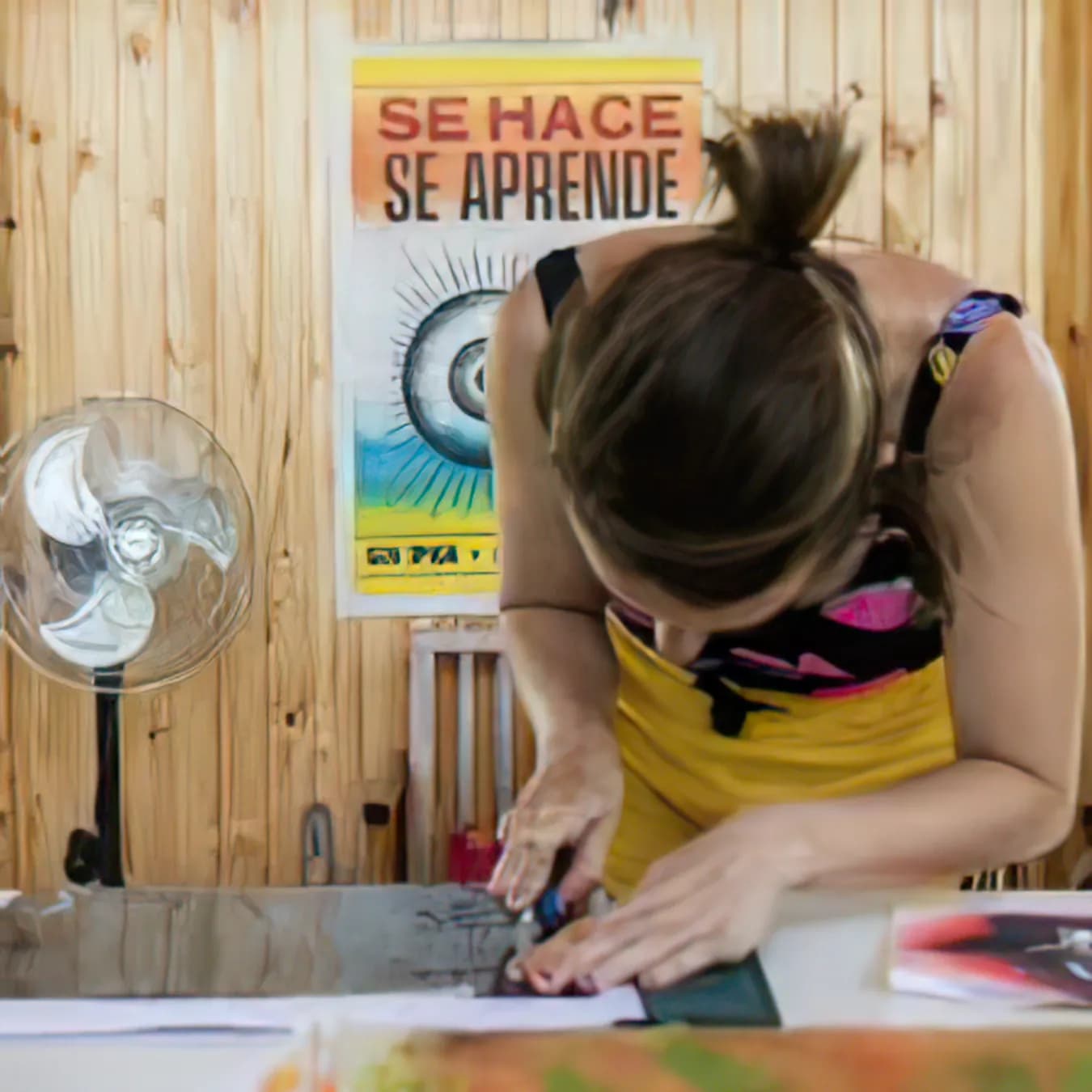 Una persona recorta papeles en un taller, frente a un afiche que dice «Se hace, se aprende»